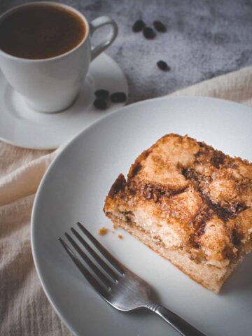 a slice of coffee cake on a plate with a fork and a cup of coffee on the side