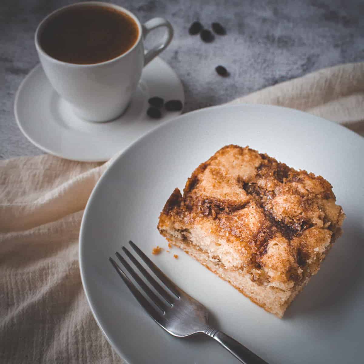a slice of coffee cake on a plate with a fork and a cup of coffee on the side