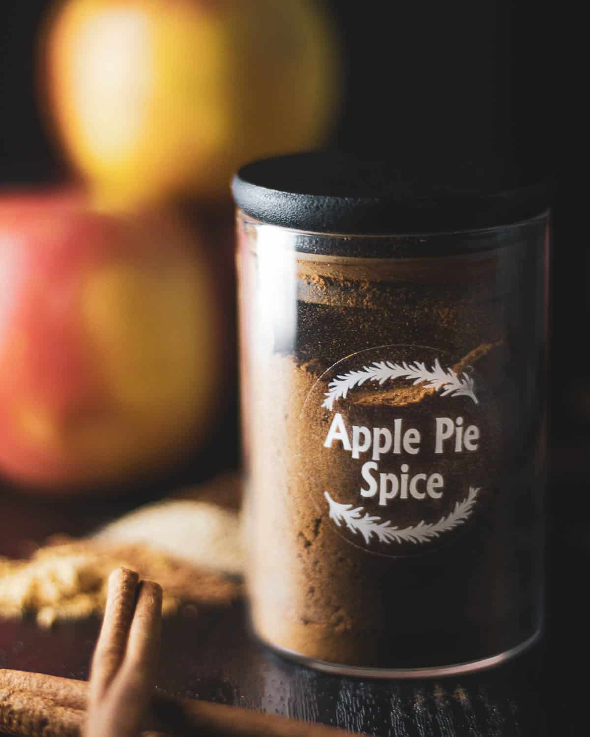 apple pie spice in a spice jar with apples and cinnamon sticks in the background