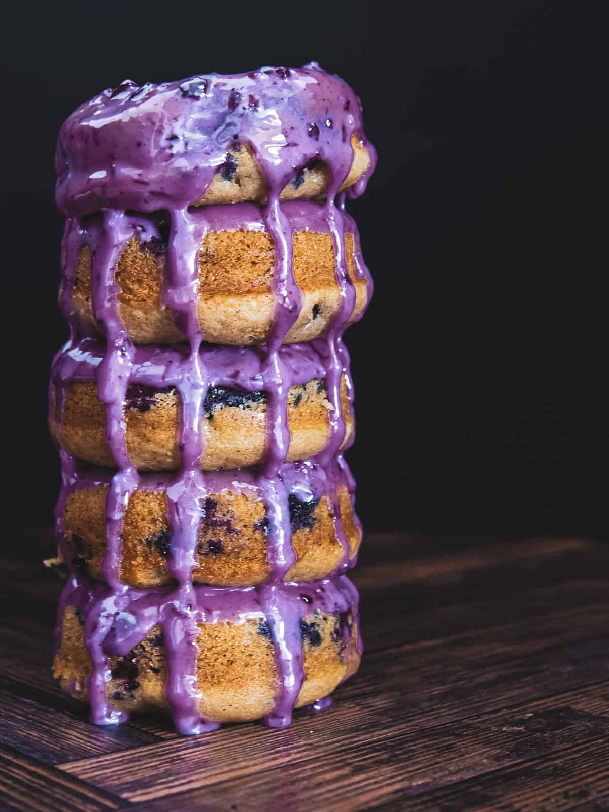 A stack of 5 blueberry donuts with blueberry glaze pouring over them.
