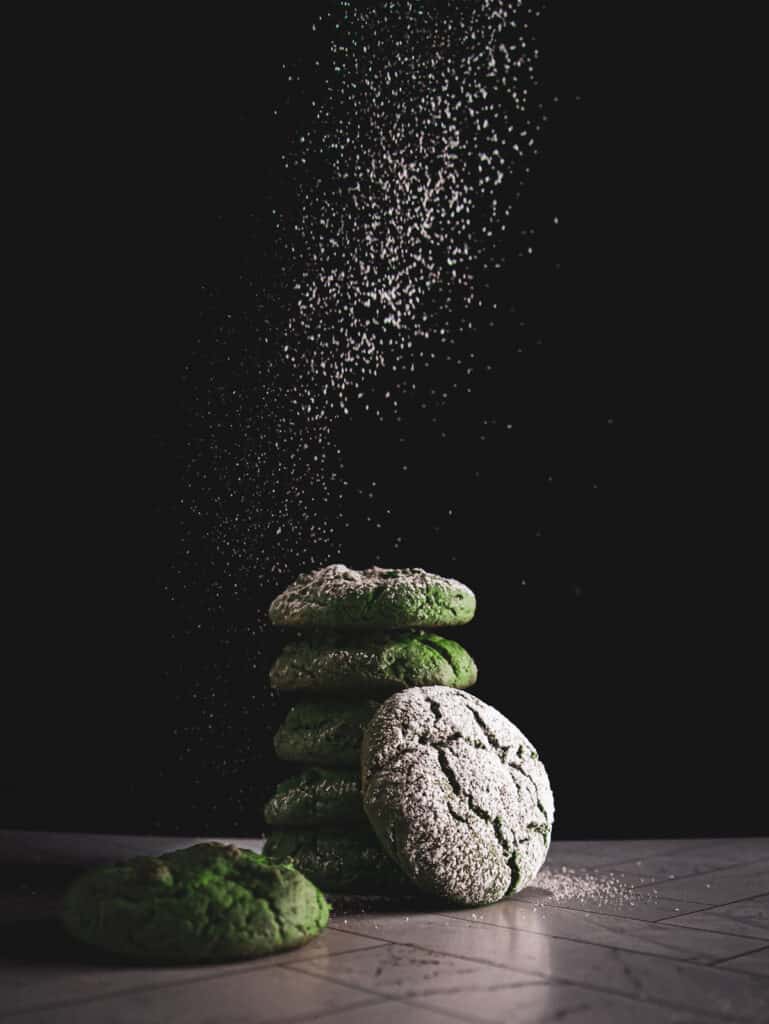 A stack of matcha crinkle cookies being dusted with powdered sugar.